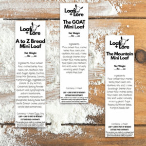 Three vertically oriented bread labels for Loaf + Lore mini loaves are displayed side by side on a floured wooden surface. Each label is white with black text and features the goat logo and “Loaf + Lore” at the top. The first label reads “A to Z Bread Mini Loaf,” the second “The GOAT Mini Loaf,” and the third “The Mountain Mini Loaf.” Each lists ingredients, net weight, and allergen information, with a small disclaimer noting compliance with Nevada cottage food regulations at the bottom. Flour dust surrounds the labels, creating a rustic, homemade bakery aesthetic.